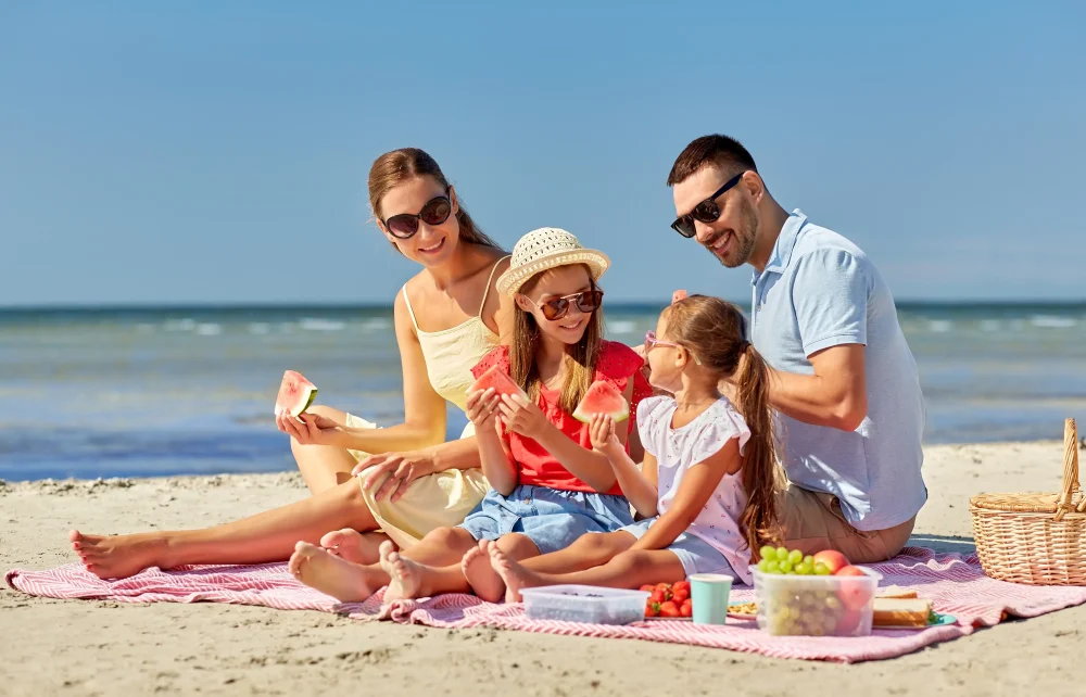 family, leisure and people concept - happy mother, father and two daughters having picnic on summer beach and eating watermelon